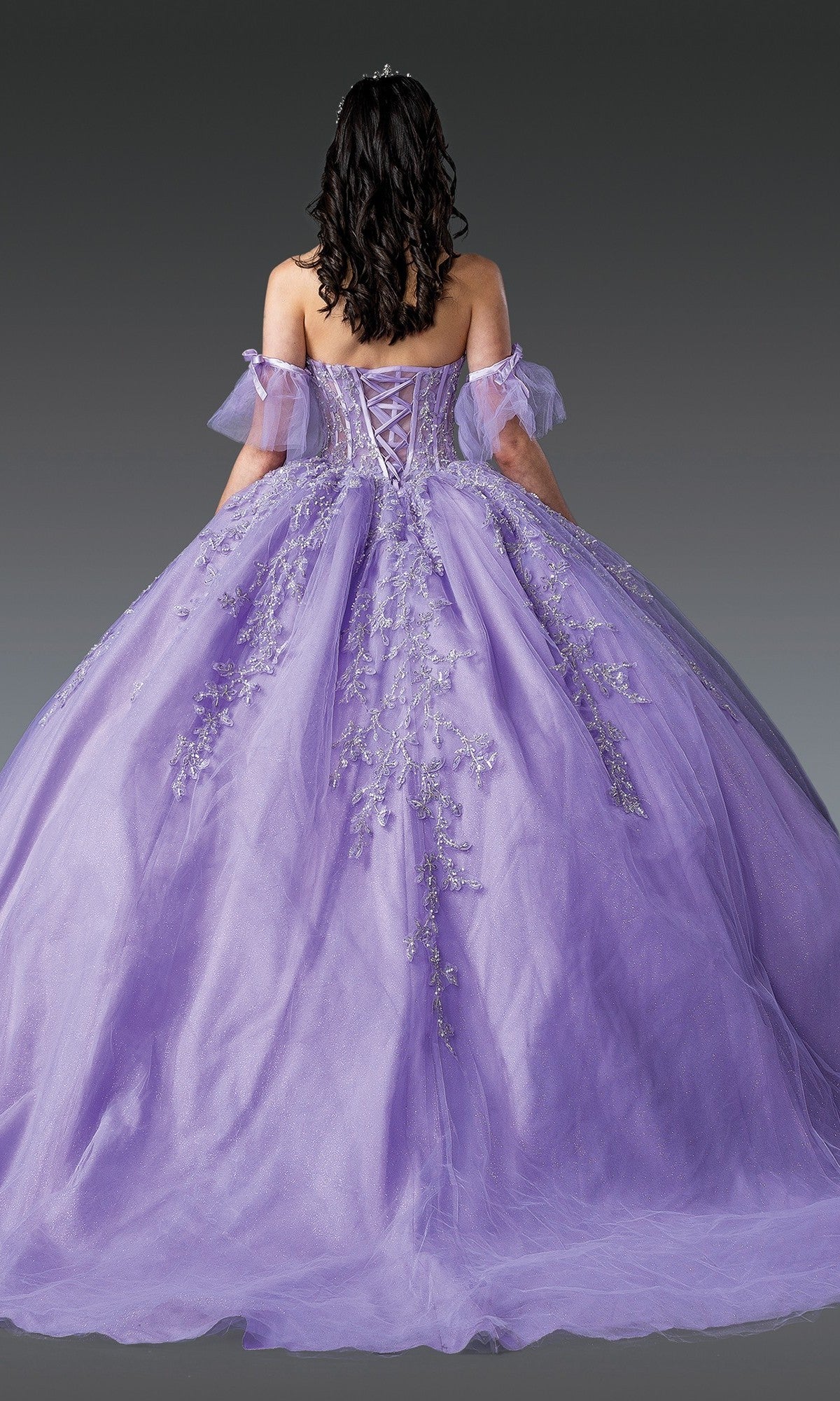 A person wearing the Quinceanera Dress 1910 By Dancing Queen, featuring a purple off-shoulder design with floral embroidery, stands facing away against a plain dark background; the ball gown skirt flows elegantly while the embellished corset bodice adds an intricate touch.