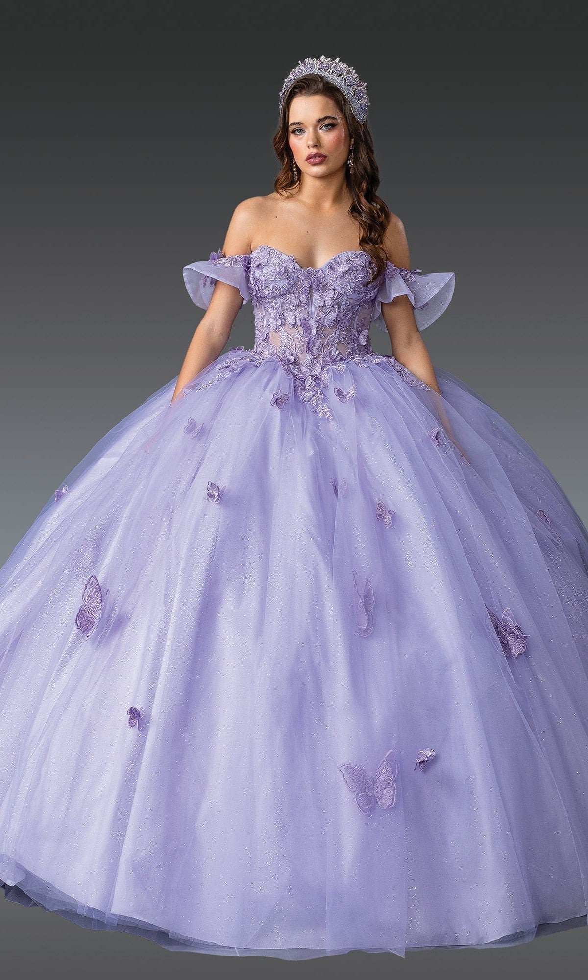 A woman stands against a gray background wearing the Quinceanera Dress 1911 by Dancing Queen in lilac, adorned with floral and butterfly appliques. The elegant ballgown features an off-the-shoulder neckline and is paired with a sparkling tiara.