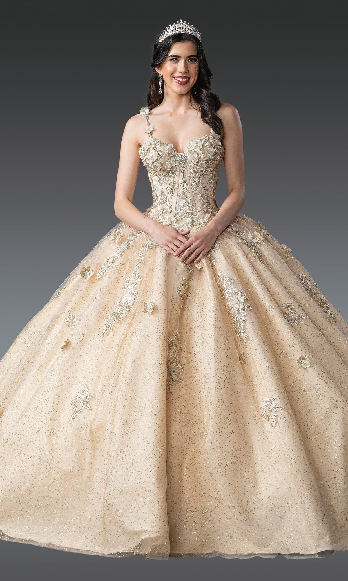 A woman stands against a gray background wearing the Quinceanera Dress 1915 By Dancing Queen, an ornate light gold glitter tulle ball gown with floral embroidery, a corset bodice, and a tiara.
