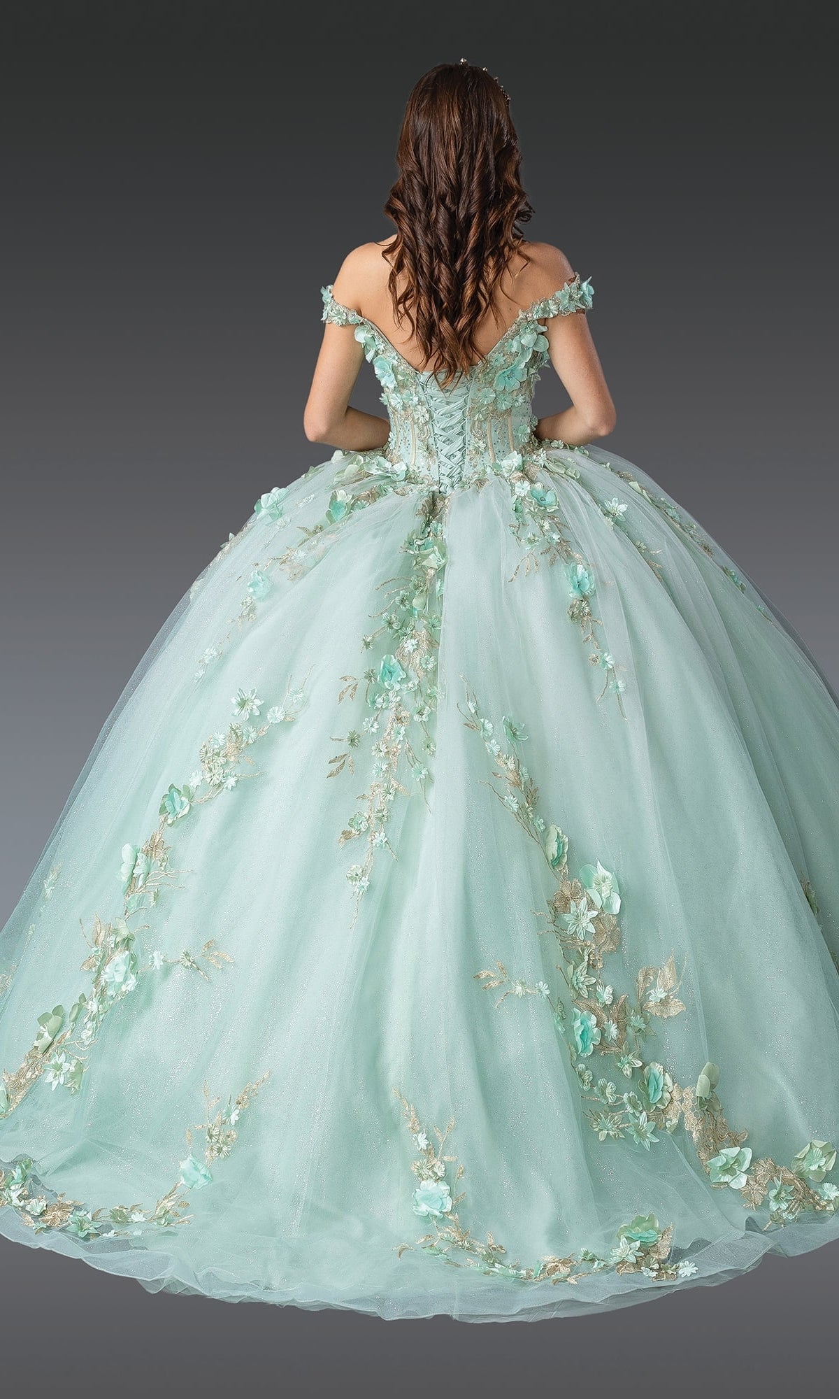 A woman faces away from the camera in the Quinceanera Dress 1918 By Dancing Queen, a mint green ball gown with floral embroidery and a corset bodice, set against a gray background.