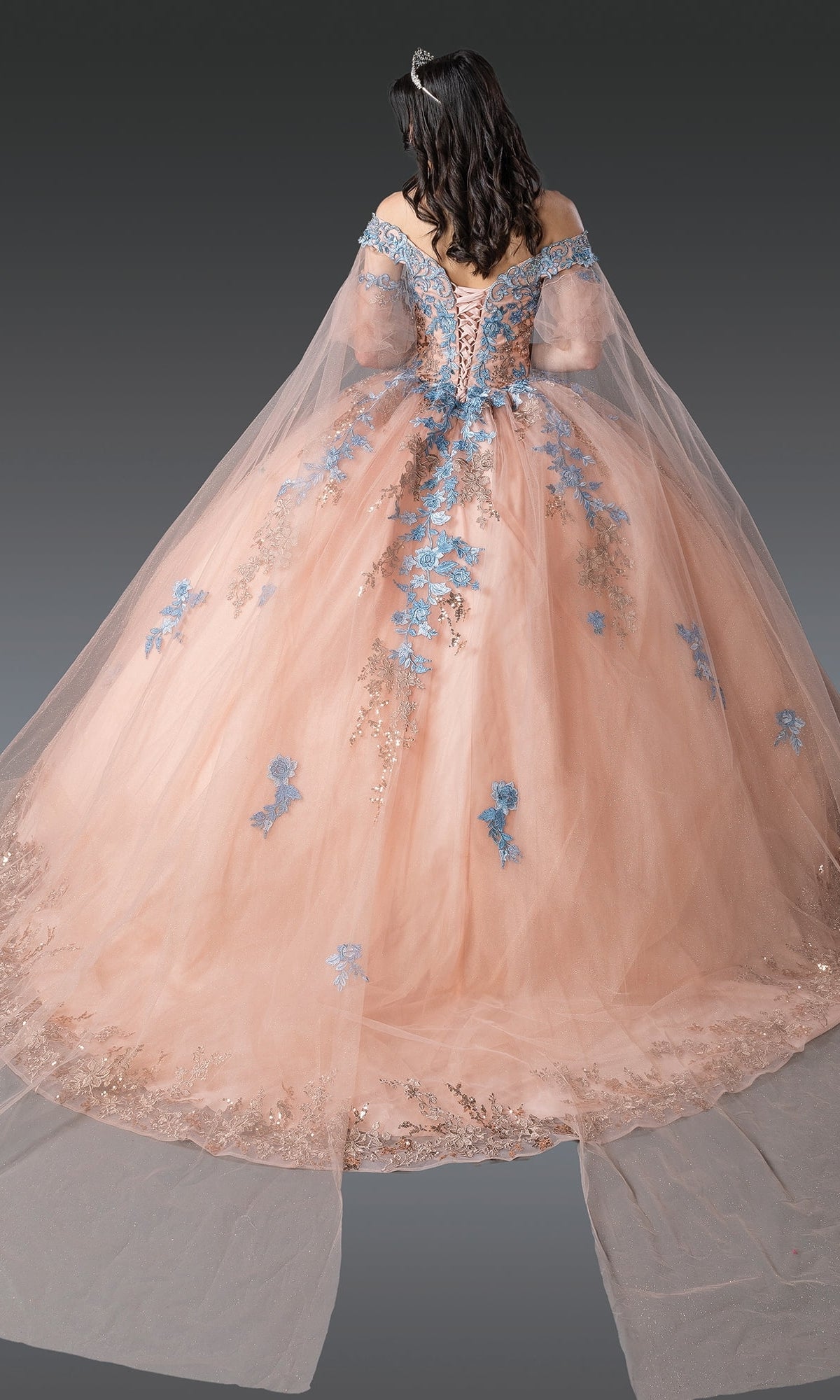 A woman stands with her back to the camera against a gray background, wearing the Quinceanera Dress 1921 By Dancing Queen—a peach ball gown with a flowing skirt, blue lace embroidery, and a sheer tulle cape.