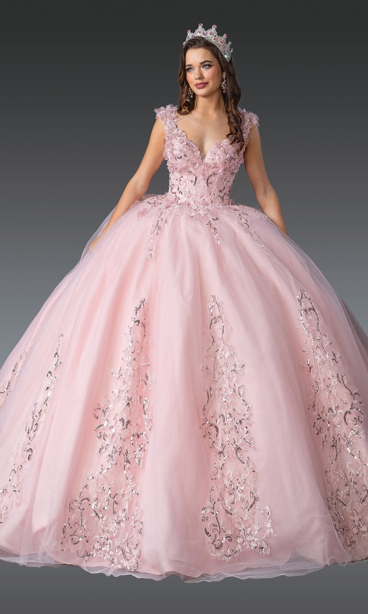 A woman in the Blush Pink Quinceanera Dress 1925 by Dancing Queen, featuring 3D flowers and a fitted bodice, poses with a tiara against a plain gray background.