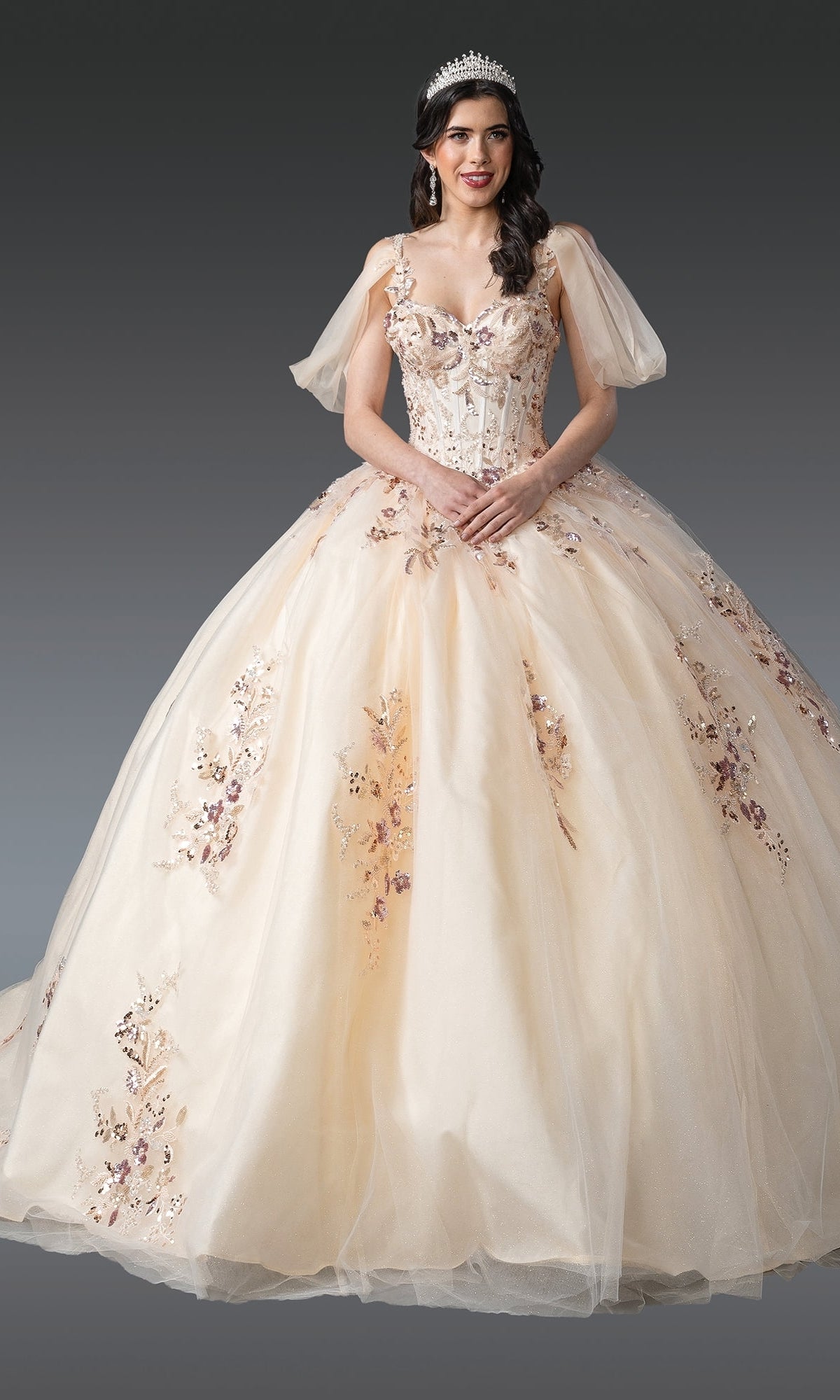 A young woman wears the Quinceanera Dress 1926 By Dancing Queen—a champagne ball gown with sequin lace embroidery, off-the-shoulder sleeves, and a beaded tiara—standing against a gray background.
