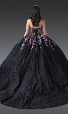 A woman stands facing away, showcasing the full skirt of the Black Quinceanera Dress 1927 By Dancing Queen with colorful floral appliqué and a tiara in her dark hair.