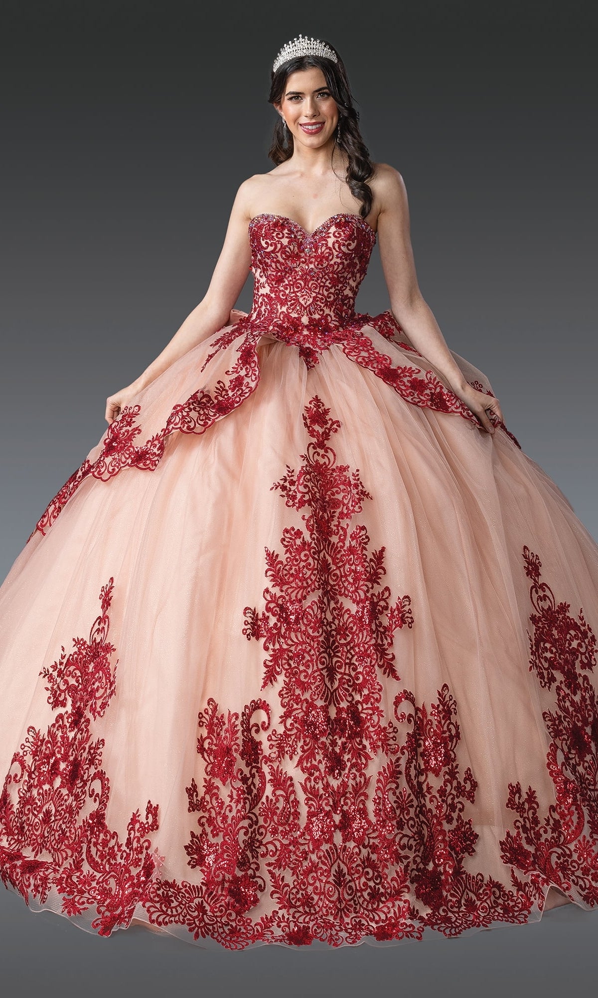 A woman wearing Quinceanera Dress 1928 By Dancing Queen, a strapless rose gold ball gown with striking burgundy red embroidery and a full skirt, poses against a gray background.
