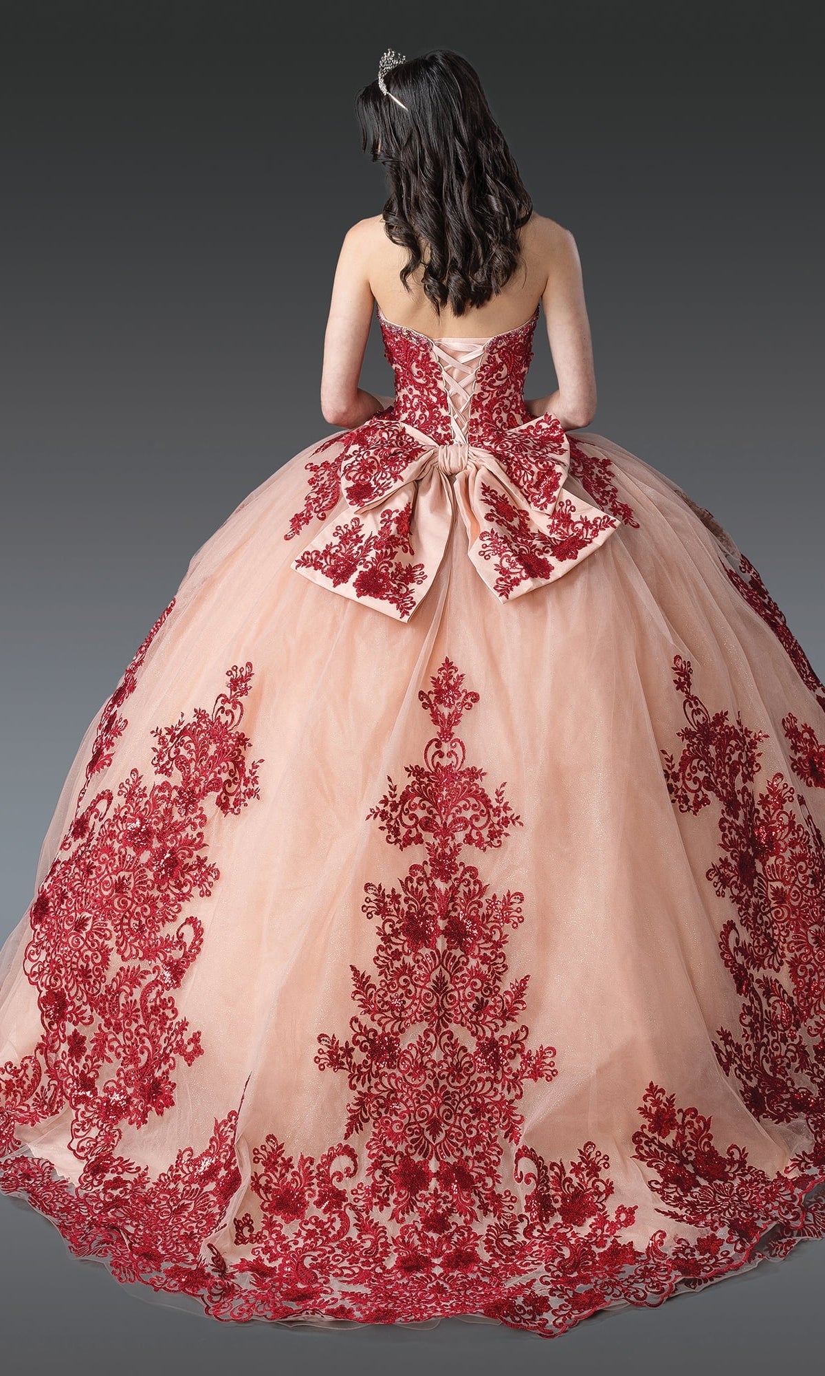 A woman wears the Quinceanera Dress 1928 By Dancing Queen, a strapless ball gown with burgundy embroidery, lace-up back, and large bow, shown from behind against a gray backdrop.