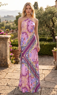 A woman stands on a stone patio wearing the Paisley Floral Printed Maxi Dress: Morgan 19309 in Purple/Ivory, featuring a long, halter neckline and vibrant purple, yellow, and blue floral patterns. Greenery and a scenic landscape in the background enhance the elegance of her floral maxi dress.