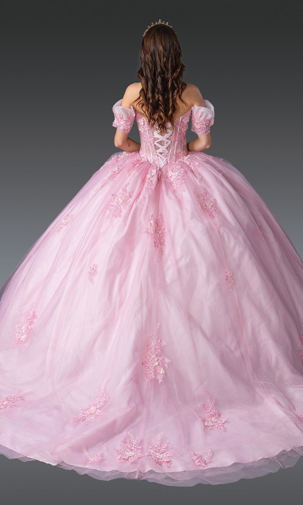 A woman with long brown hair is seen from the back wearing Quinceanera Dress 1932 By Dancing Queen, a pink off-the-shoulder gown with floral appliqués and a corset bodice, set against a plain gray background.
