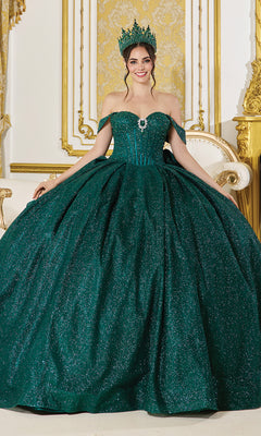 A woman in the Quinceanera Dress 1937 by Dancing Queen—a sparkling emerald green glitter ball gown with off-the-shoulder straps—wears a matching crown and stands in an ornate gold and white room.
