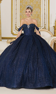 A woman stands in an ornate room wearing the Quinceanera Dress 1937 By Dancing Queen—a dark blue, off-the-shoulder ball gown with a full sparkling skirt—paired with a jeweled necklace and tiara.