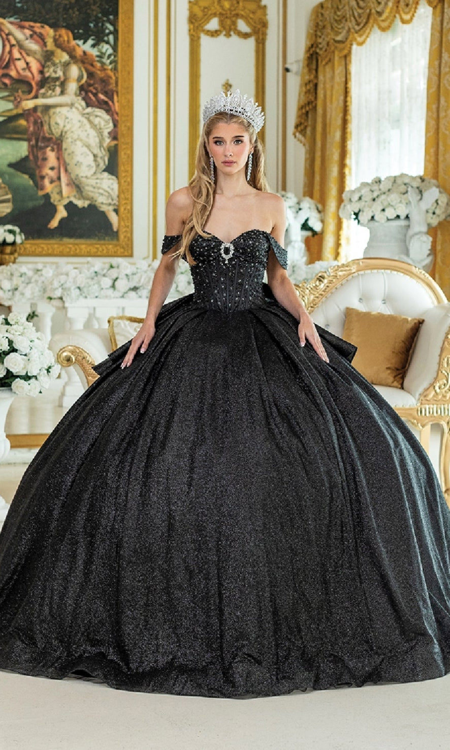 A woman wears the Quinceanera Dress 1937 By Dancing Queen, a black glitter ball gown with off-the-shoulder straps and a tiara, standing in an ornate room with gold decor, white flowers, and classical artwork.
