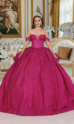A woman in the Quinceanera Dress 1937 By Dancing Queen, a sparkling bright pink ball gown with off-the-shoulder straps, wears a silver tiara and stands in an ornate room decorated with white flowers and gold accents.