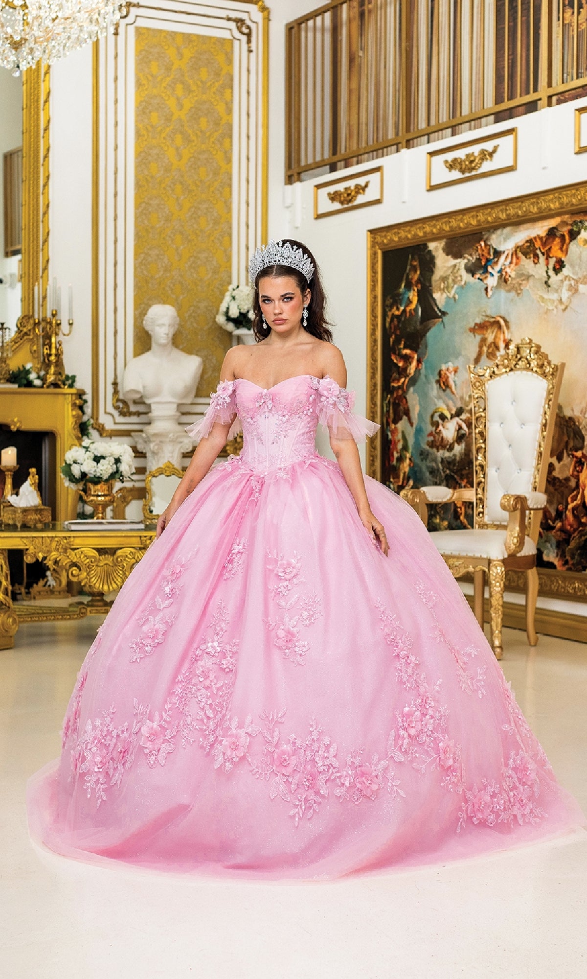 A woman wearing the Quinceanera Dress 1944 By Dancing Queen, featuring a pink corset bodice and floral details, stands in an ornate room with gold accents, a white throne chair, and classical artwork.