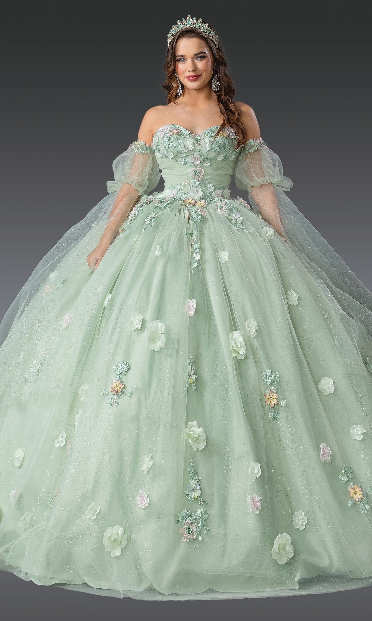 A woman in the Quinceanera Dress 1945 by Dancing Queen, featuring sage green off-shoulder puffy sleeves, 3D flowers, a grand ball gown skirt, and a tiara, stands against a plain gradient background.