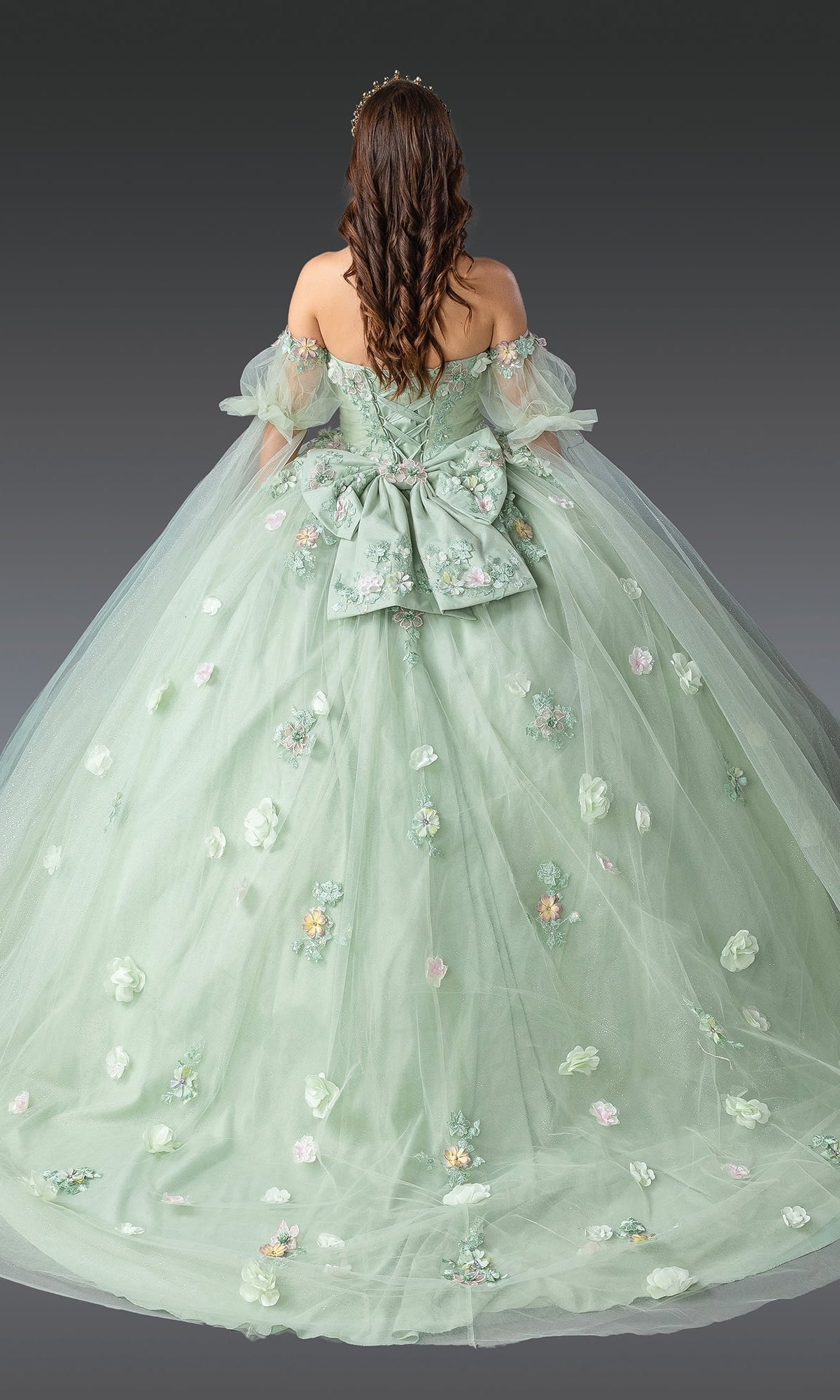 A woman wears the Quinceanera Dress 1945 by Dancing Queen in sage green, featuring 3D flowers, a large bow, an off-the-shoulder neckline, and a voluminous ball gown skirt as she stands facing away.