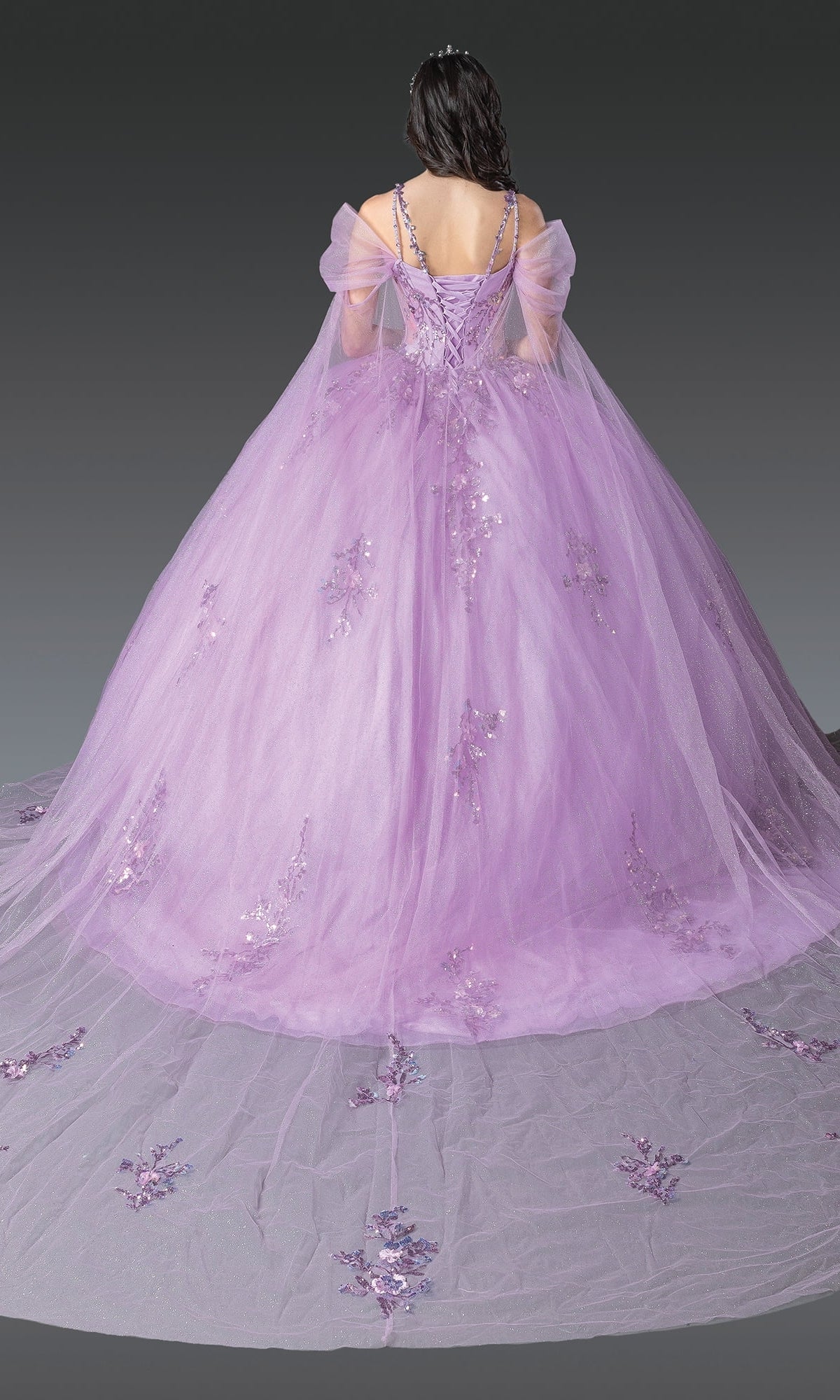 A person wearing the Quinceanera Dress 1947 By Dancing Queen, a floor-length lavender gown with a lace embroidered bodice, sheer off-shoulder sleeves, and floral embellishments on the skirt, viewed from the back.