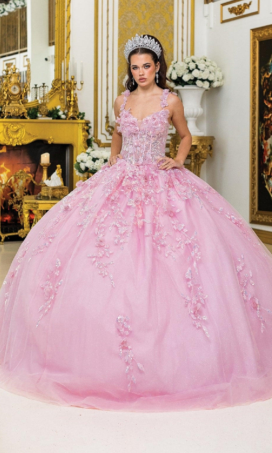 A woman in the Quinceanera Dress 1948 By Dancing Queen, featuring a sparkling pink 3D flower design with a corset bodice and ball gown skirt, stands in an ornate room adorned with gold decor and white flowers.