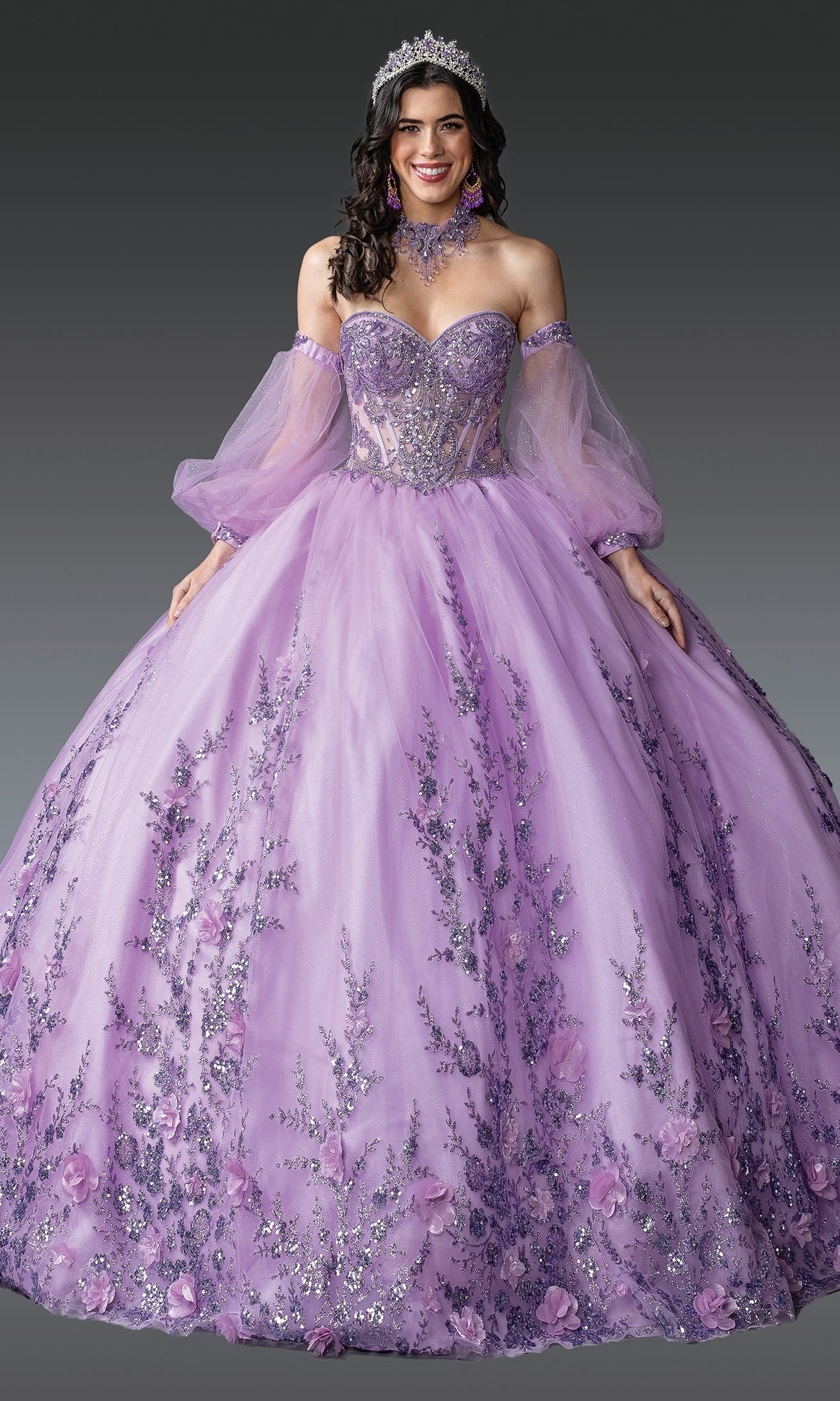 A woman smiles against a gray background, wearing Quinceanera Dress 1953 By Dancing Queen in lilac purple with sparkly floral embellishments, sheer sleeves, and a tiara.