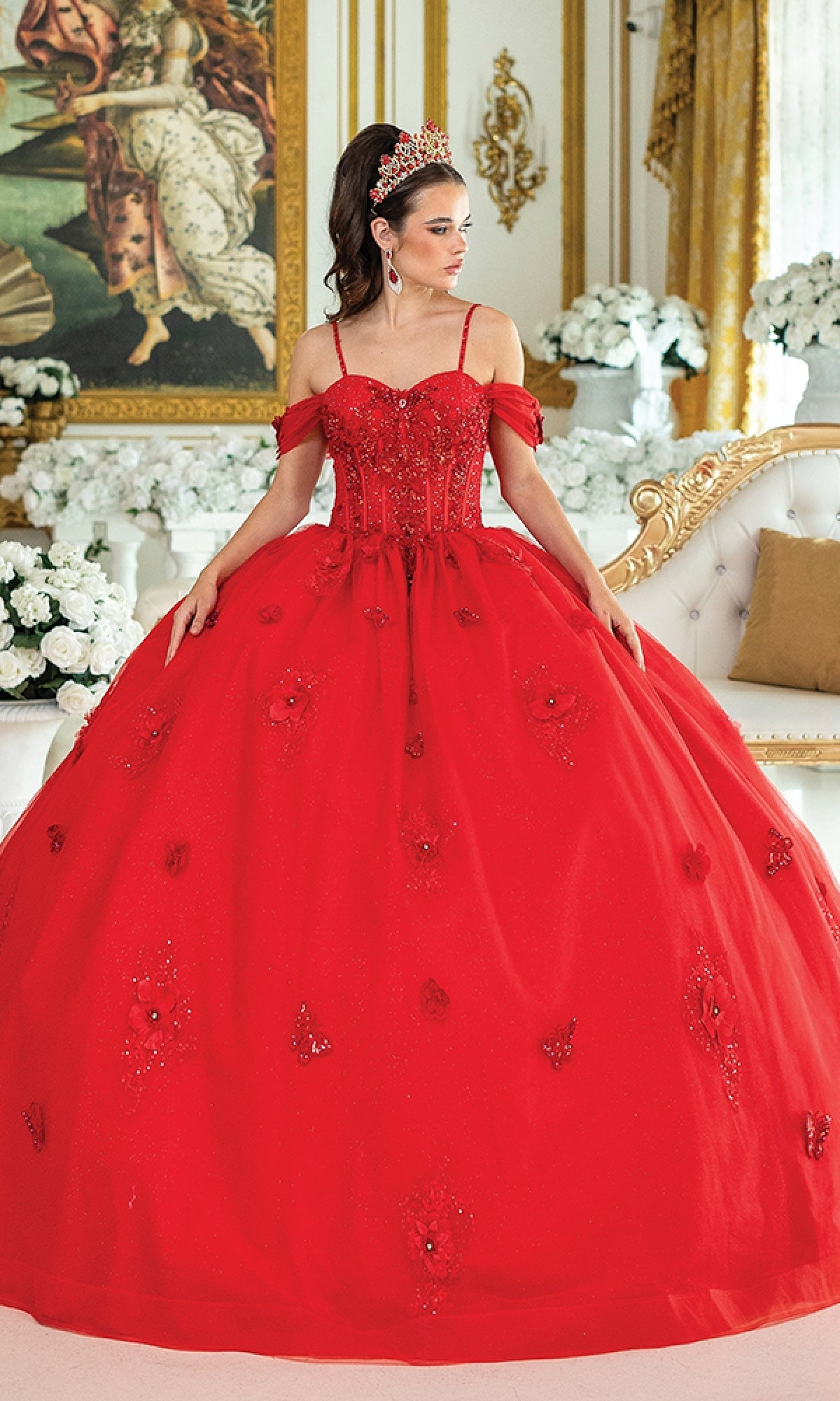 A woman wearing the Quinceanera Dress 1954 By Dancing Queen—a glitter tulle ball gown with lace corset bodice and tiara—stands in an opulent room adorned with white roses and gold accents, gazing to her left.
