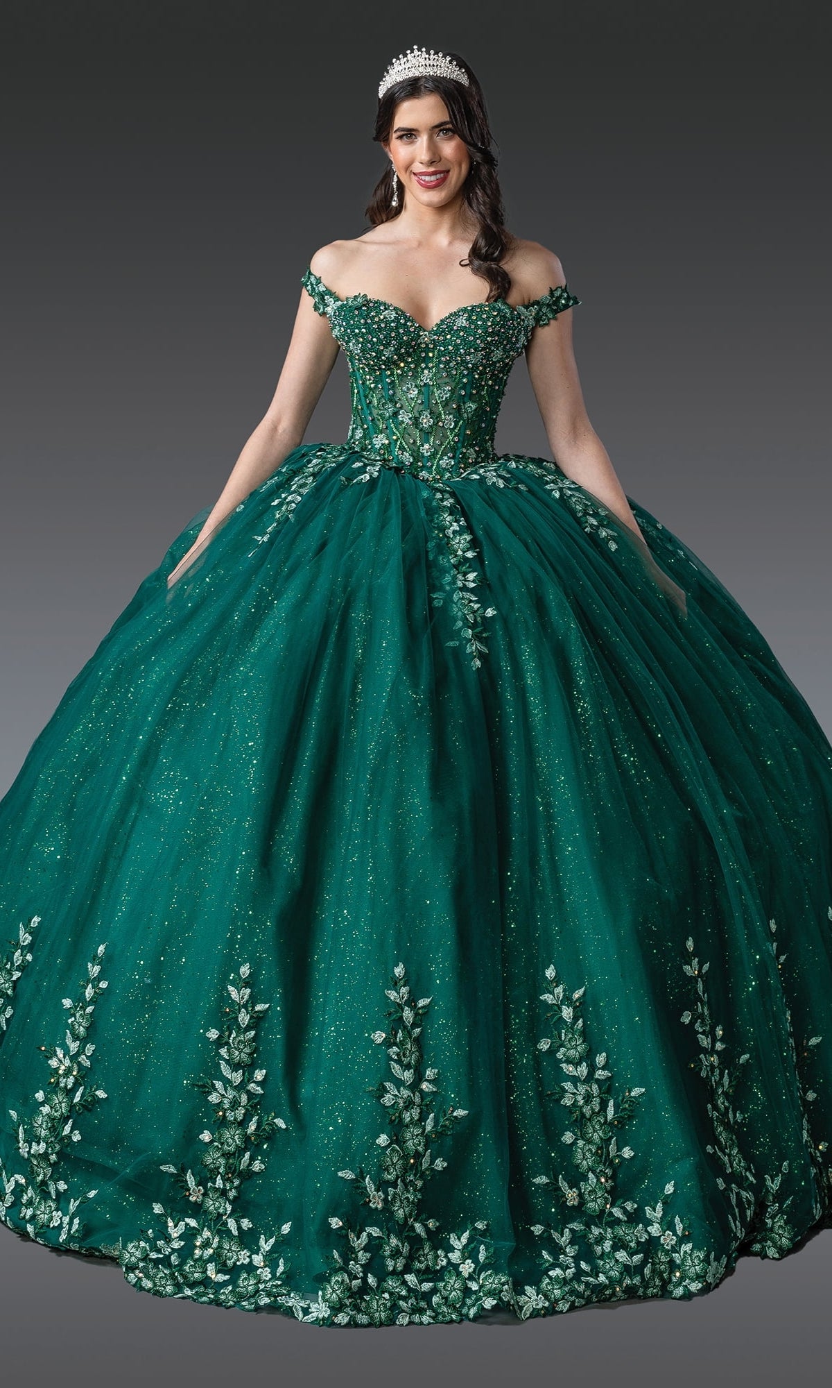 A woman poses against a dark background wearing the Quinceanera Dress 1955 By Dancing Queen—an emerald green off-the-shoulder gown with floral lace embroidery, sparkly details, and a tiara.