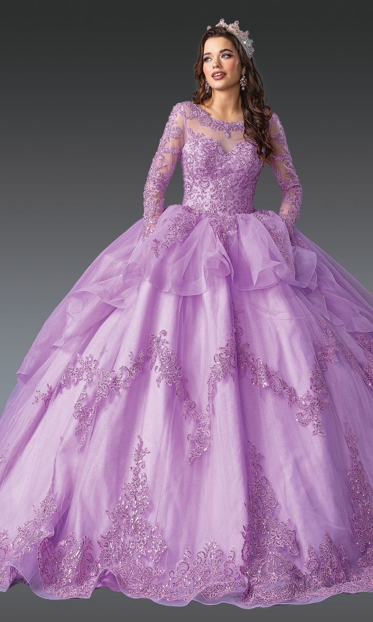 A woman poses against a dark background in the Quinceanera Dress 1956 By Dancing Queen, featuring ornate sequin lace embroidery, long sleeves, a full lilac skirt, and a sparkling jeweled tiara.