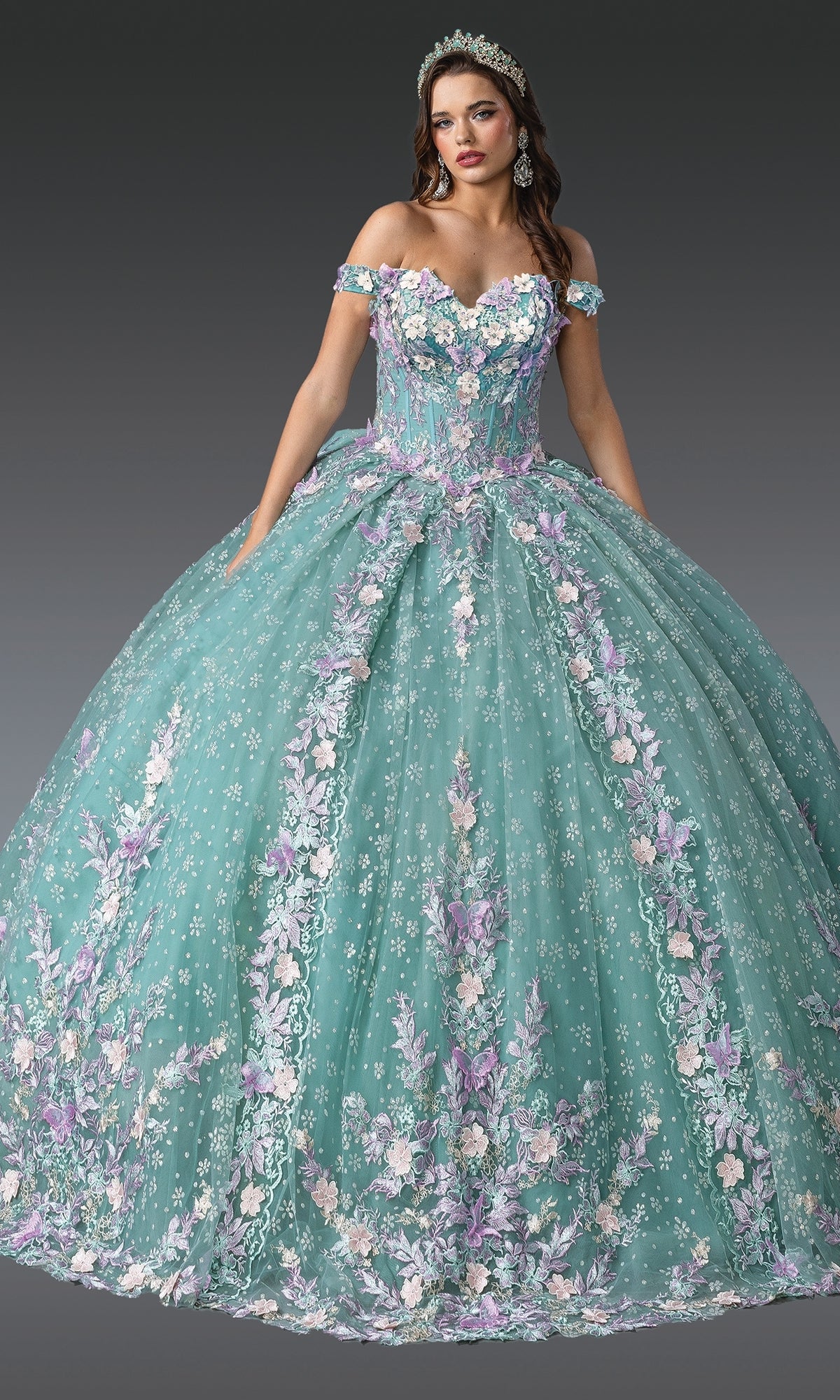 A woman in the Quinceanera Dress 1962 By Dancing Queen, a large pastel teal ball gown with floral embroidery, off-the-shoulder sleeves, and a matching tiara stands against a plain dark background.