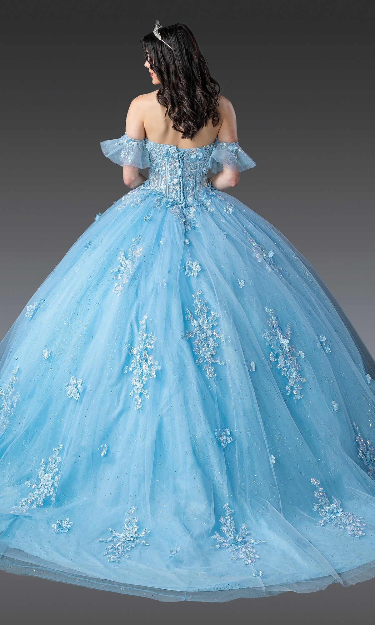 A woman with long dark hair, wearing a silver tiara and the Blue Quinceanera Dress 1964 By Dancing Queen with floral embroidery, stands facing away against a gray background.