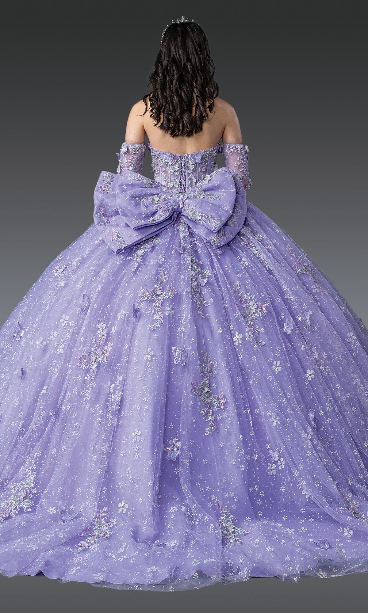 A woman faces away, wearing the Quinceanera Dress 1965 By Dancing Queen—a floor-length lavender gown with floral embroidery, off-the-shoulder sleeves, and a large bow at the back.