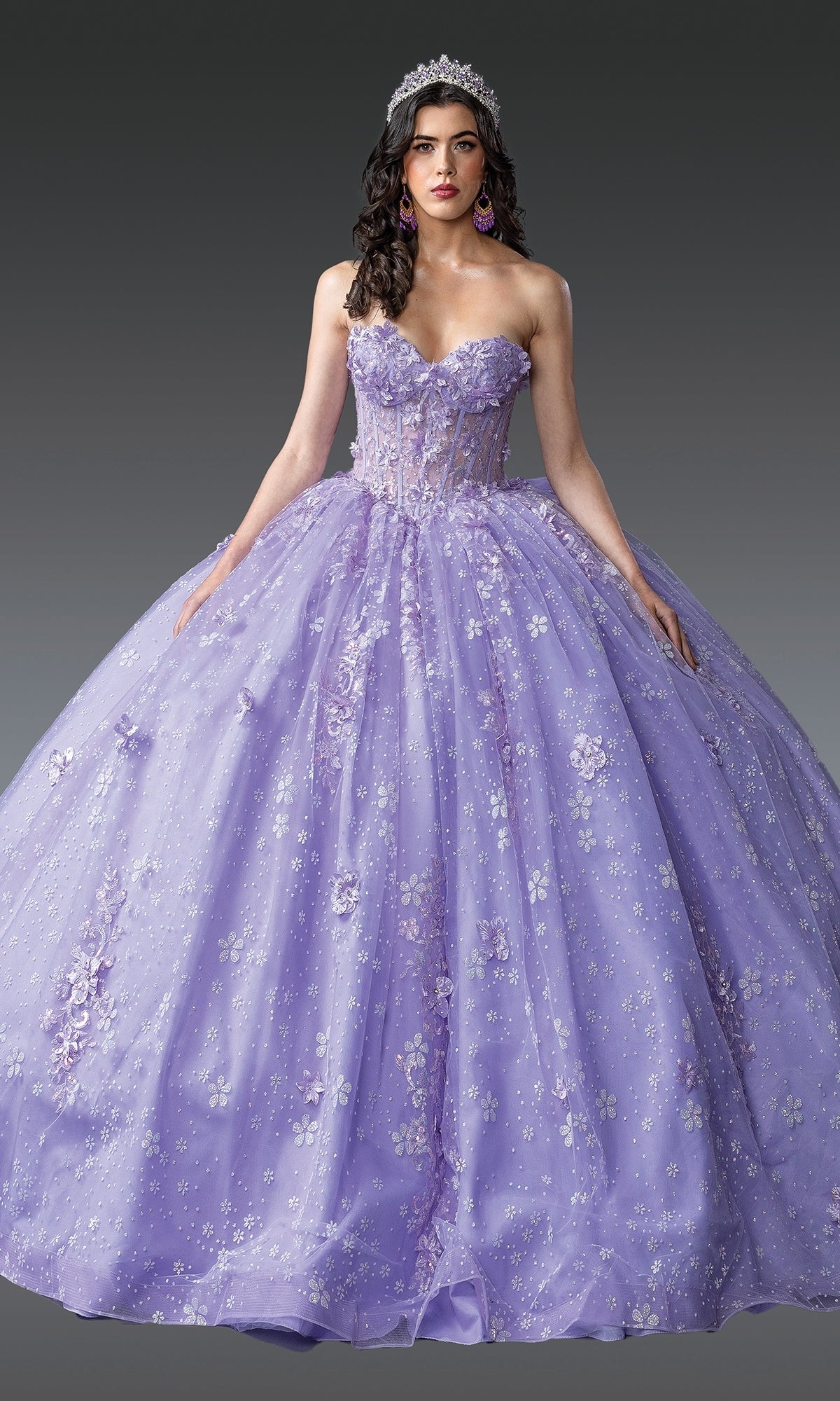 A woman wearing the Quinceanera Dress 1967 By Dancing Queen, featuring a strapless sweetheart neckline and floral embellishments in lilac purple, with a tiara, stands against a plain gray background.