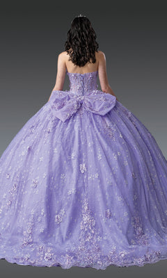 A person with wavy dark hair wears the Quinceanera Dress 1967 By Dancing Queen—a strapless, lavender ball gown with a sweetheart neckline, floral embellishments, and a large back bow—standing against a gray background.