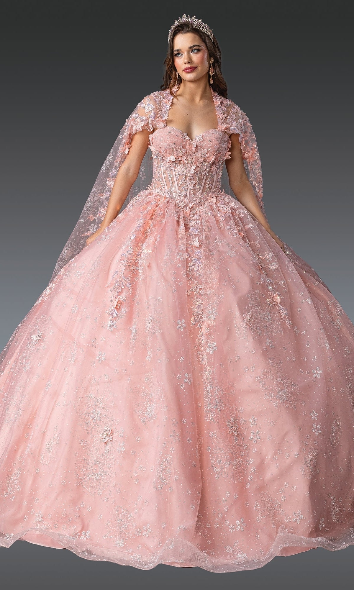A woman stands against a dark background wearing the Quinceanera Dress 1970 By Dancing Queen—an embellished light pink ball gown with 3D flowers, floral details, a matching cape, and a tiara.