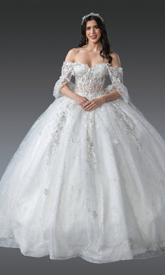 A woman in the Quinceanera Dress 1970 By Dancing Queen, an off-the-shoulder white gown with 3D flowers and a full tulle ball skirt, wears a sparkling headpiece while standing against a plain gray background.