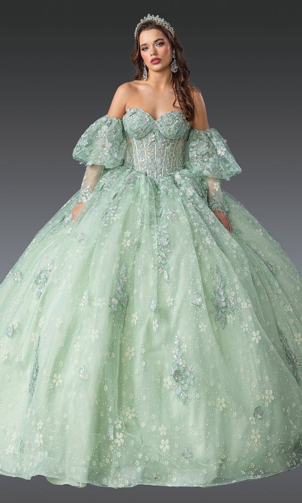 A woman in the Quinceanera Dress 1973 By Dancing Queen, featuring a light green off-the-shoulder corset bodice, floral embroidery, and puff sleeves, stands against a plain dark background.