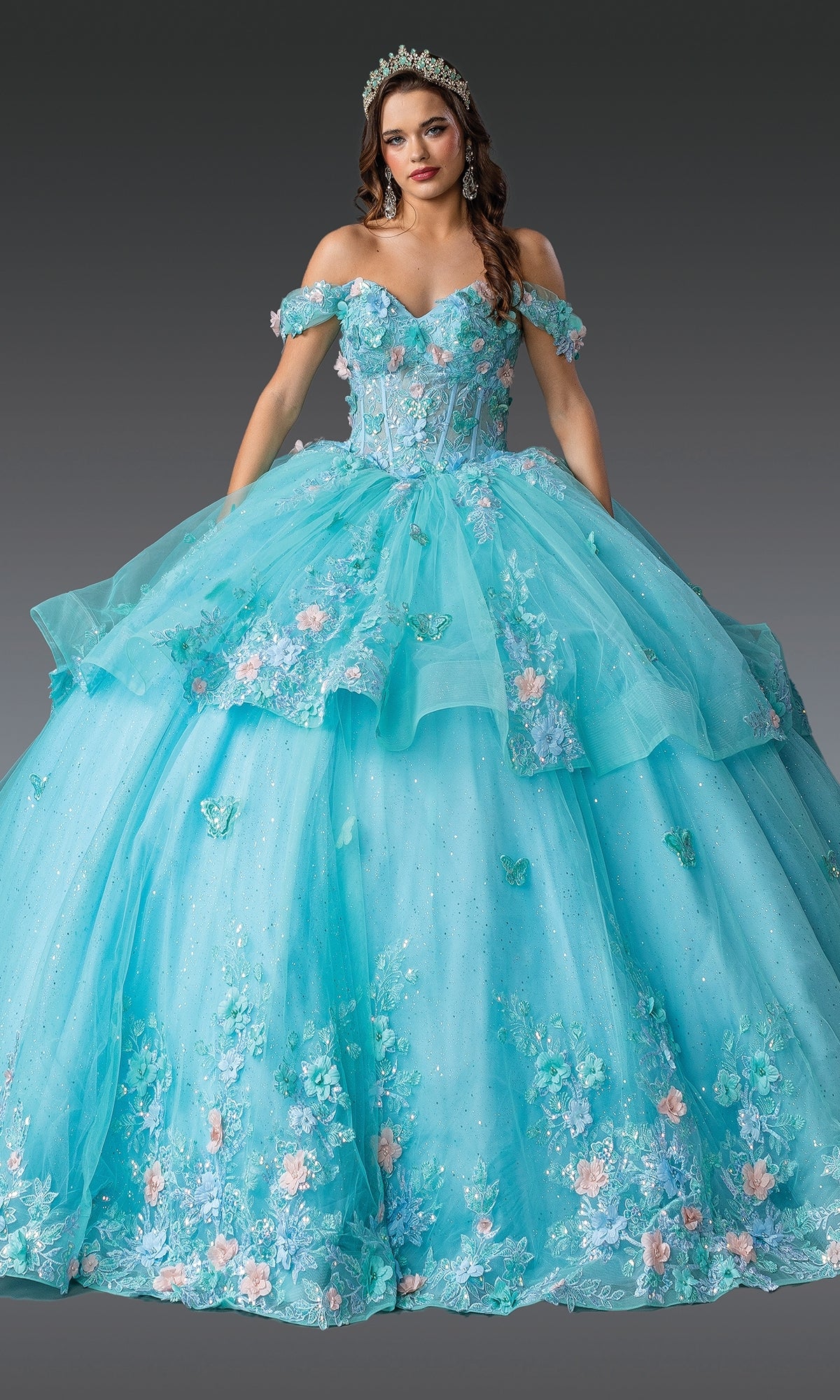 A woman in the Quinceanera Dress 1974 By Dancing Queen—a light blue, off-the-shoulder gown with floral appliqué and a layered tulle skirt—wears a tiara as she stands against a plain gray background.