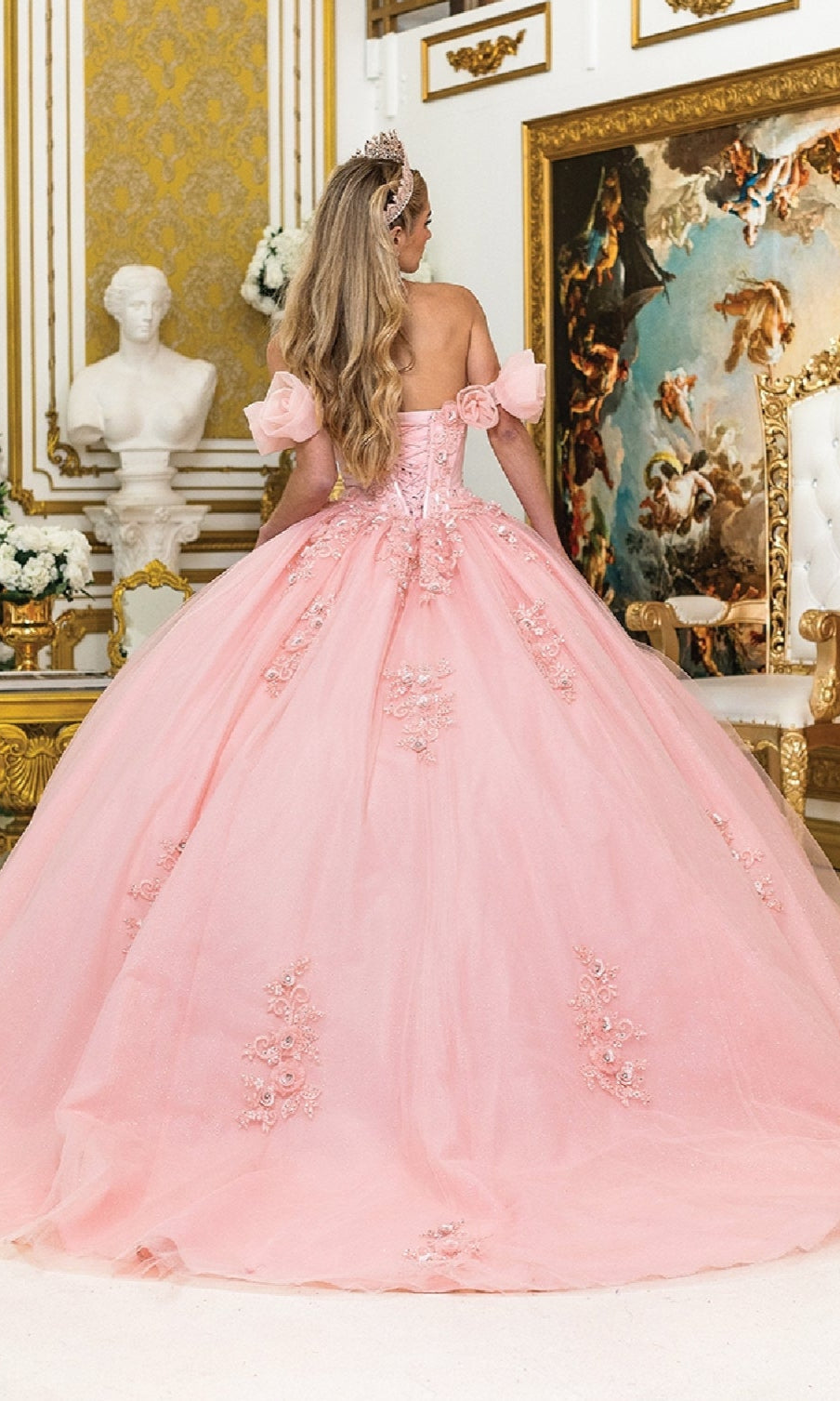 A woman wearing the Quinceanera Dress 1976 by Dancing Queen, featuring a pink corset bodice, stands with her back to the camera in an ornate room with gold accents and classical decor.