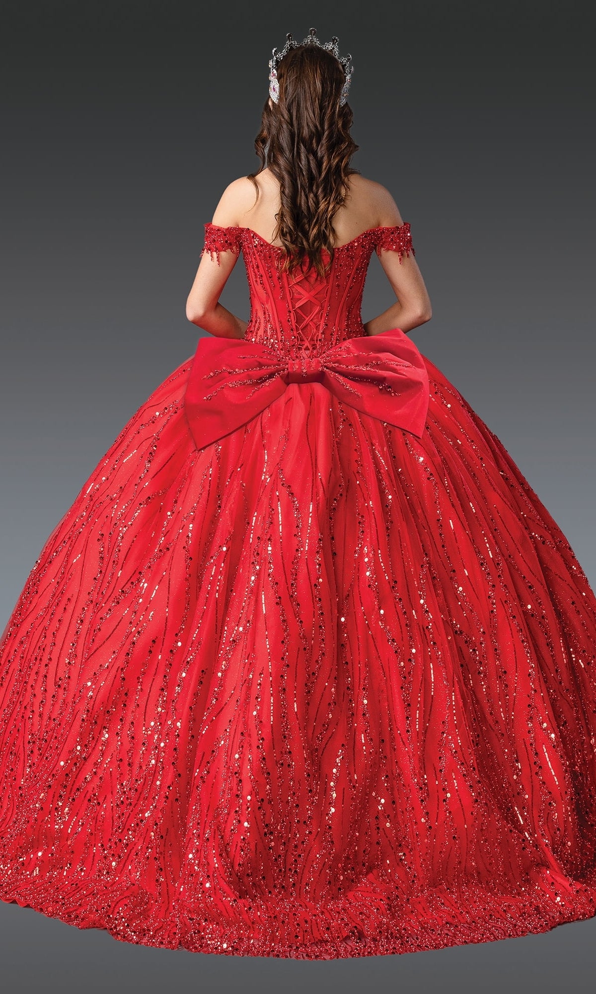 A person in the Long Corset Quinceañera Dress: Dancing Queen 1982, a floor-length sequin-embellished ball gown with a large back bow and jeweled hair accessory, is facing away from the camera.
