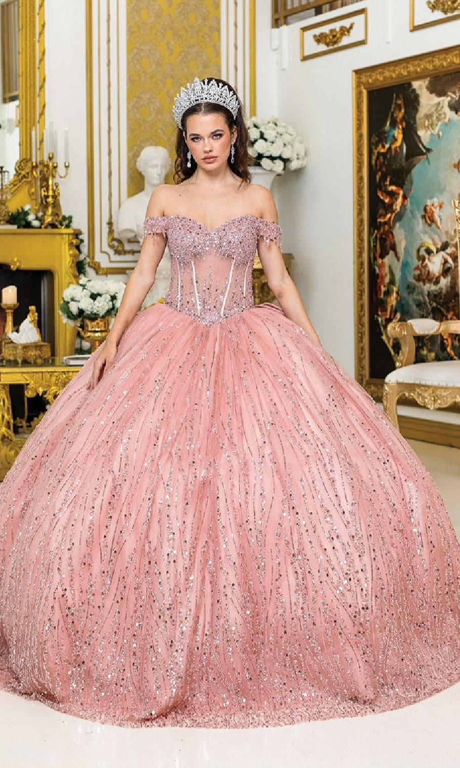 A woman wears the Long Corset Quinceañera Dress: Dancing Queen 1982 in an ornate room with gold decor, white flowers, and classical artwork in the background.