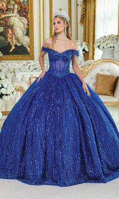 A woman wearing the Long Corset Quinceañera Dress: Dancing Queen 1982 stands in an ornate, gold-accented room with white florals, her sequin-embellished ball gown and jeweled crown sparkling in the light.