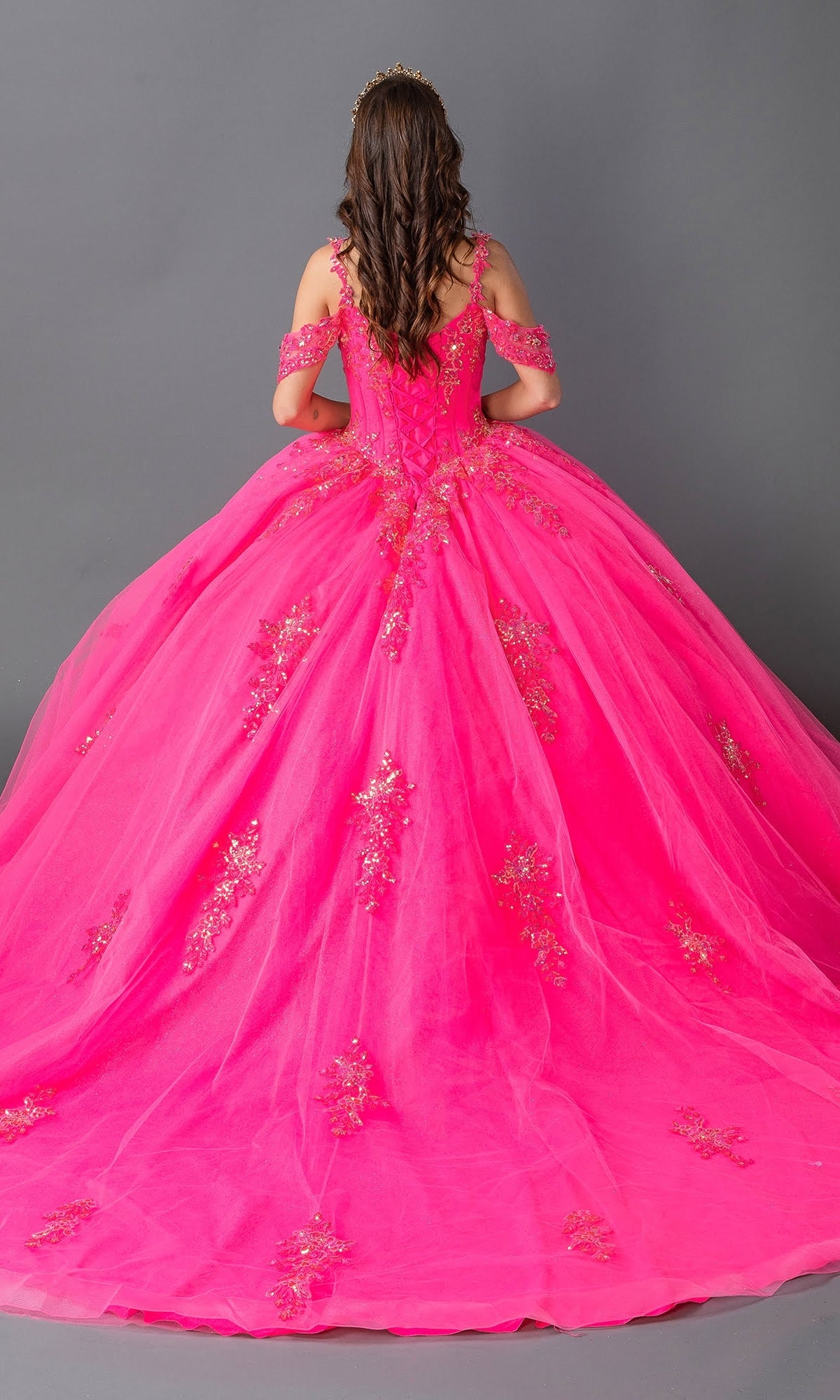 A young woman faces away from the camera in the Hot Pink Quinceanera Dress 1983 By Dancing Queen, featuring gold floral lace embroidery and a corset bodice, set against a gray background.