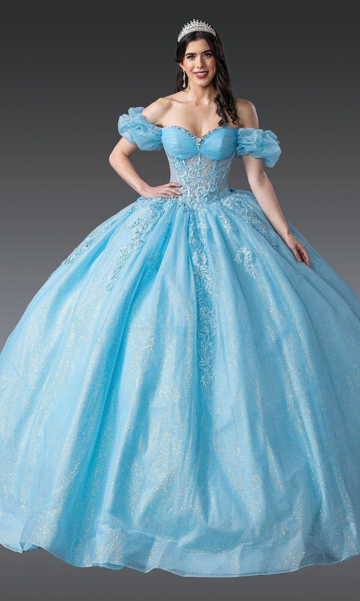 A woman wears the Quinceanera Dress 1985 By Dancing Queen, a blue off-the-shoulder ball gown with a lace embroidered corset bodice and a tiara, standing against a plain gray background.