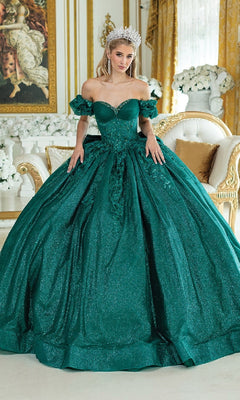 A woman wearing the Quinceanera Dress 1985 By Dancing Queen, a sparkling emerald green ball gown with a corset bodice and tiara, stands in an ornate room adorned with gold decor, white flowers, and elegant furniture.