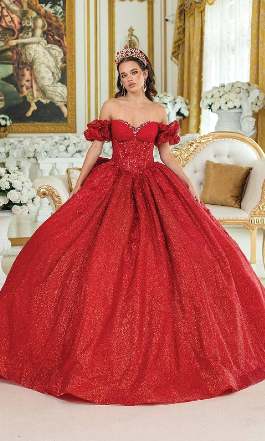 A woman in the Quinceanera Dress 1985 By Dancing Queen, a sparkling red ball gown with a corset bodice and crown, stands in an ornate room decorated with white flowers, gold accents, and a classical painting.