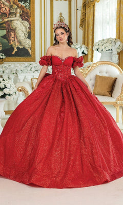 A woman in the Quinceanera Dress 1985 By Dancing Queen, a sparkling red ball gown with a corset bodice and crown, stands in an ornate room decorated with white flowers, gold accents, and a classical painting.