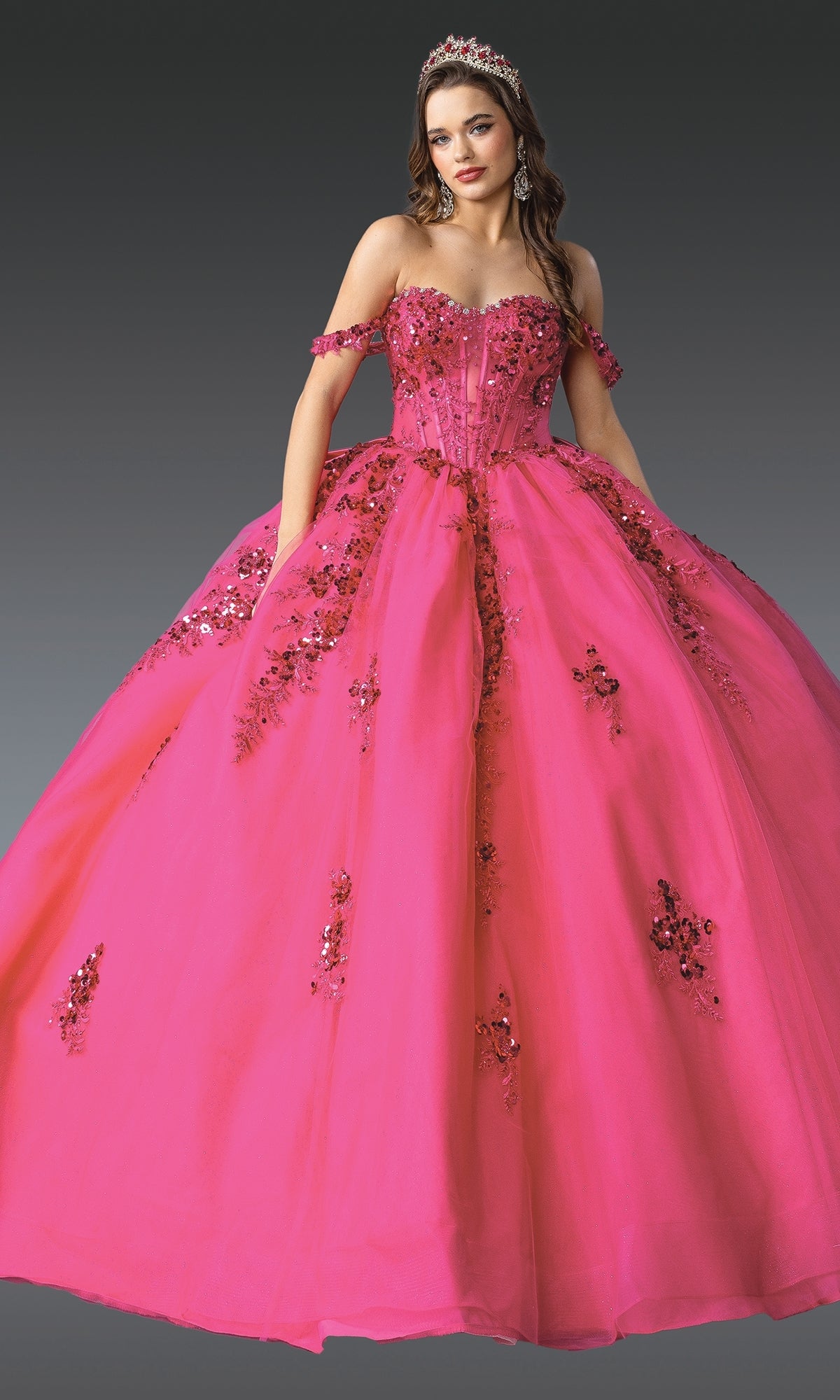 A woman in the Quinceanera Dress 1986 By Dancing Queen, featuring a bright pink hue, corset bodice, floral embellishments, tiara, and off-the-shoulder sleeves, stands against a plain gray background.