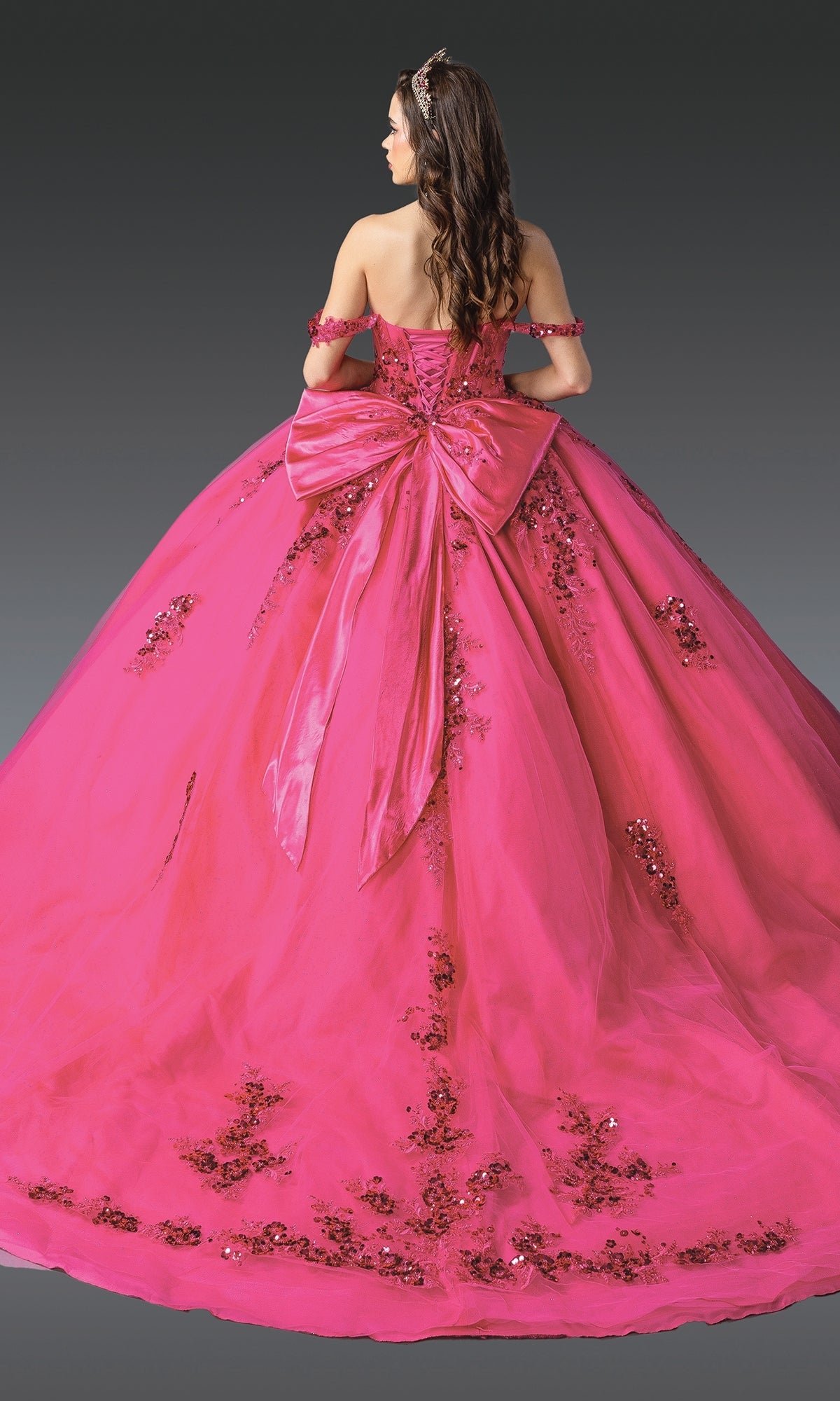A woman wearing the Quinceanera Dress 1986 by Dancing Queen, featuring a bright pink hue, corset bodice, off-the-shoulder sleeves, sequin embellishments, and a large back bow, stands against a dark background.