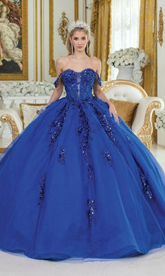A woman in the Quinceanera Dress 1986 by Dancing Queen—a sparkling blue glitter tulle ball gown with a corset bodice and tiara—stands in an ornate room adorned with gold accents and white flowers.