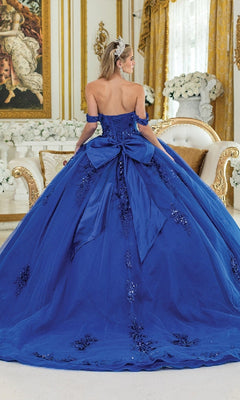 In an ornate room, a woman faces away from the camera wearing the Quinceanera Dress 1986 By Dancing Queen—royal blue with an off-the-shoulder corset bodice, a large back bow, and dark floral embellishments.