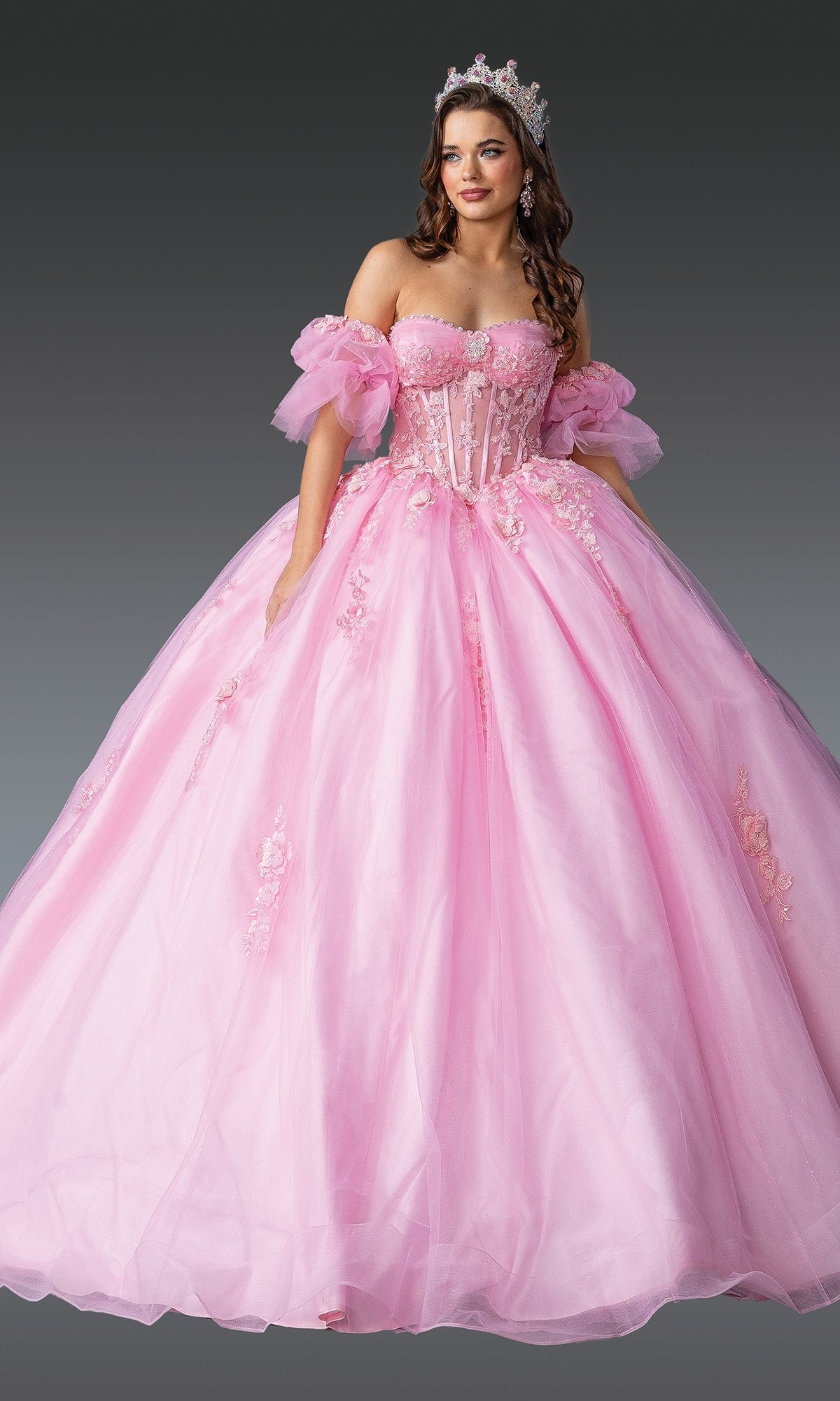 A woman in the Pink Quinceanera Dress 1988 By Dancing Queen, featuring lace embroidery, floral details, and a tiara, poses against a gray background in an elegant off-the-shoulder ball gown.