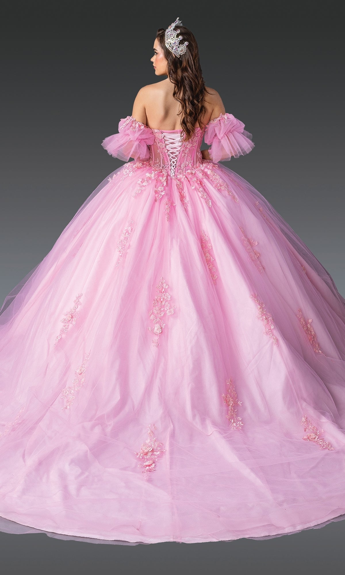 A woman faces away, wearing the Pink Quinceanera Dress 1988 By Dancing Queen—an off-the-shoulder pink gown with lace embroidery and a full skirt—paired with a decorative headpiece in her long, wavy hair.