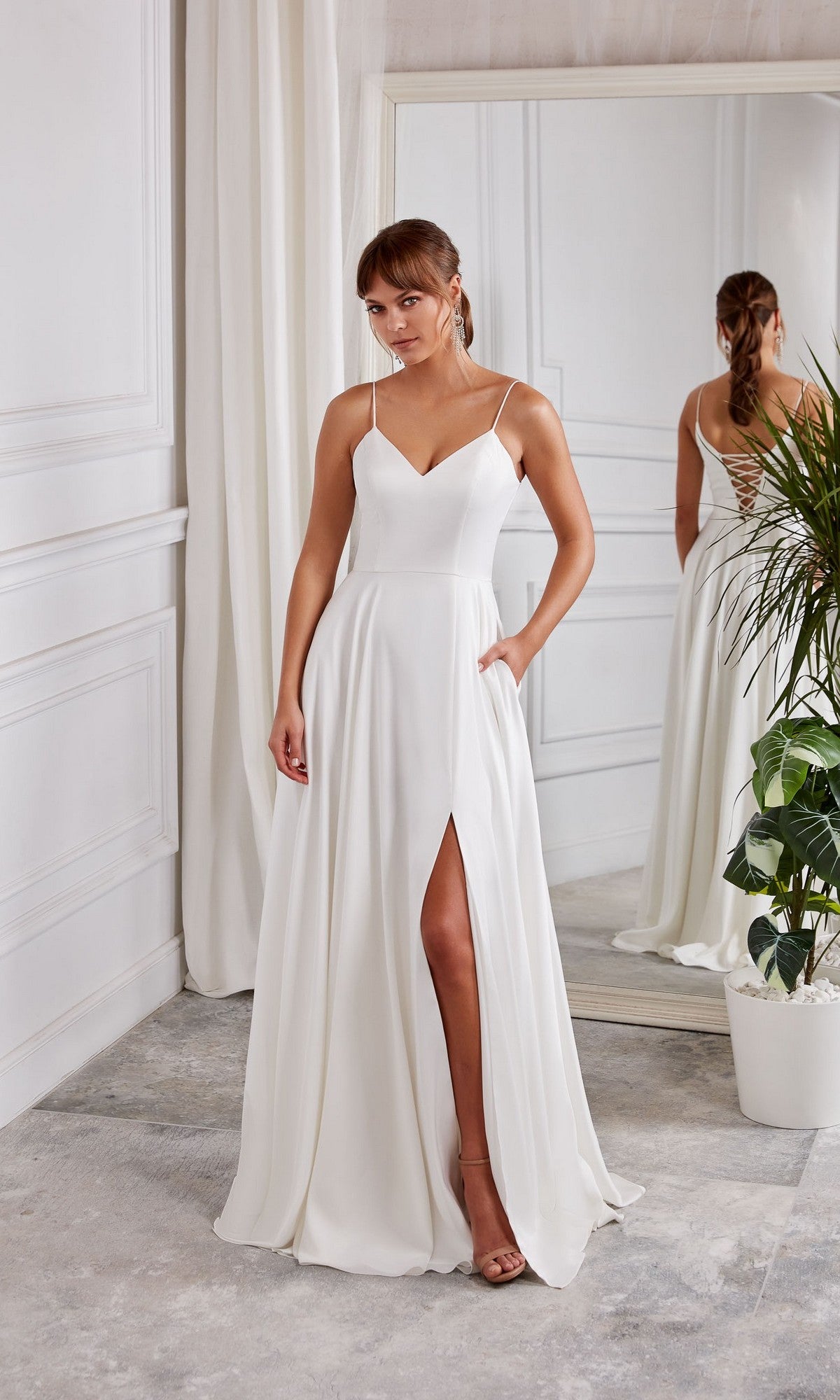 A woman wearing the Aerie Couture 2001 Long White Dress in Diamond White, featuring an a-line skirt and thin straps, stands indoors near a mirror and a potted plant.